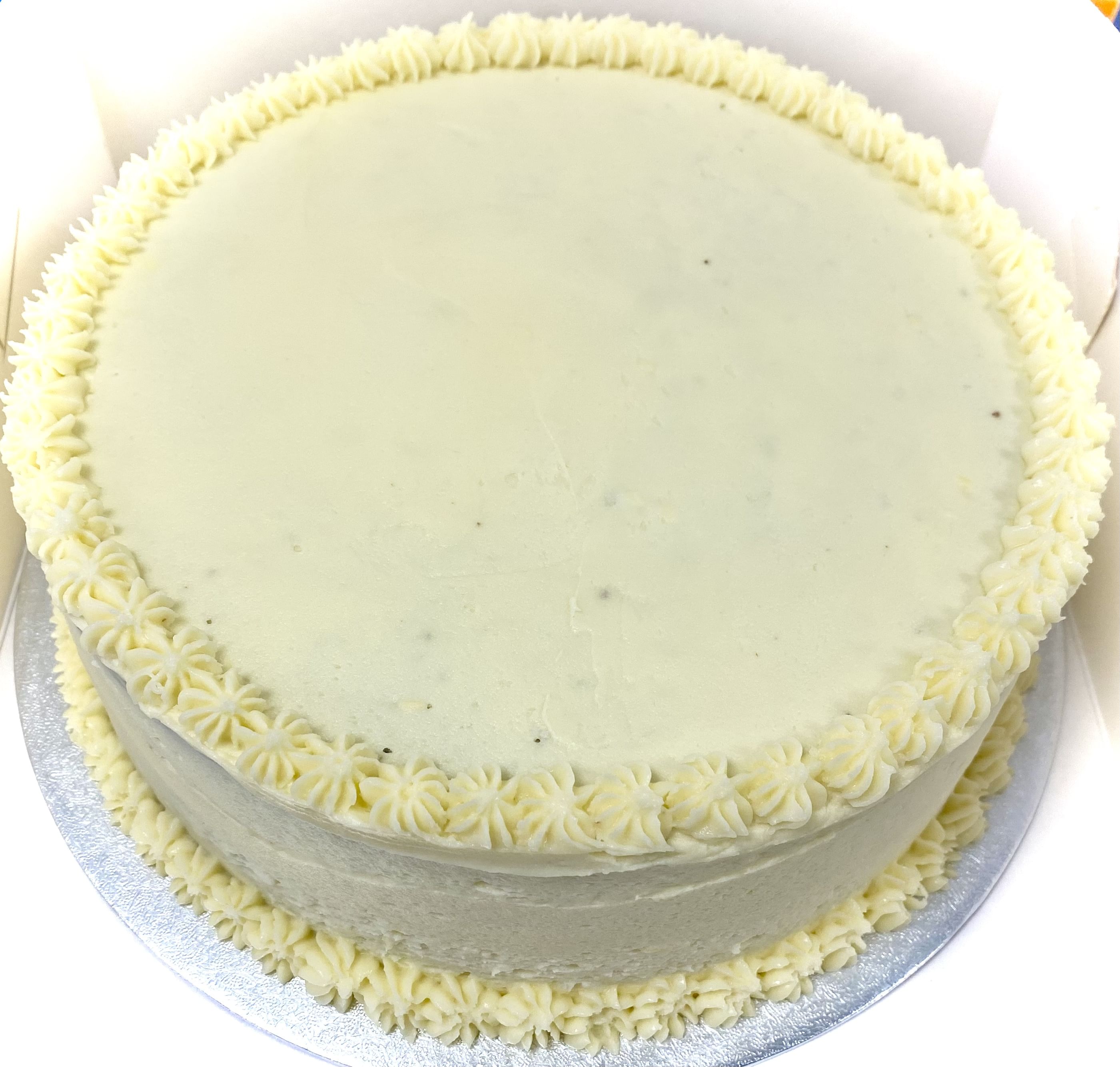 A round, plain white frosted cake with a simple design. The cake has small star-shaped frosting details along the top edge and the base. It is presented in a white box, with a silver cake board beneath it. The cake appears smooth and uniform in texture.